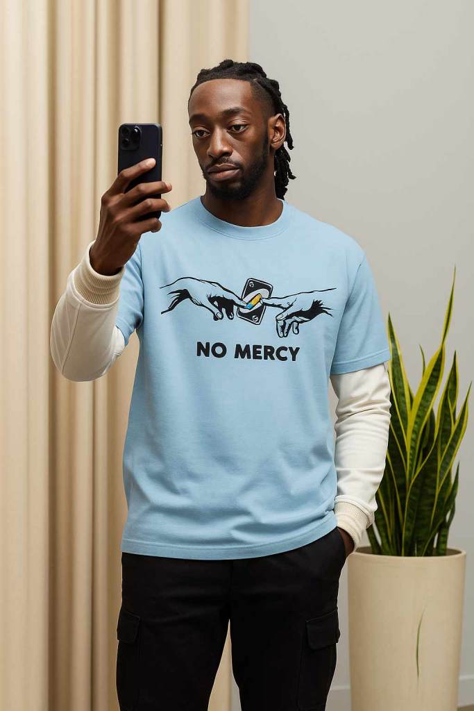 A person holding a smartphone while wearing a light blue t-shirt with the words 'NO MERCY' printed on it, set against a neutral background with indoor plants.
